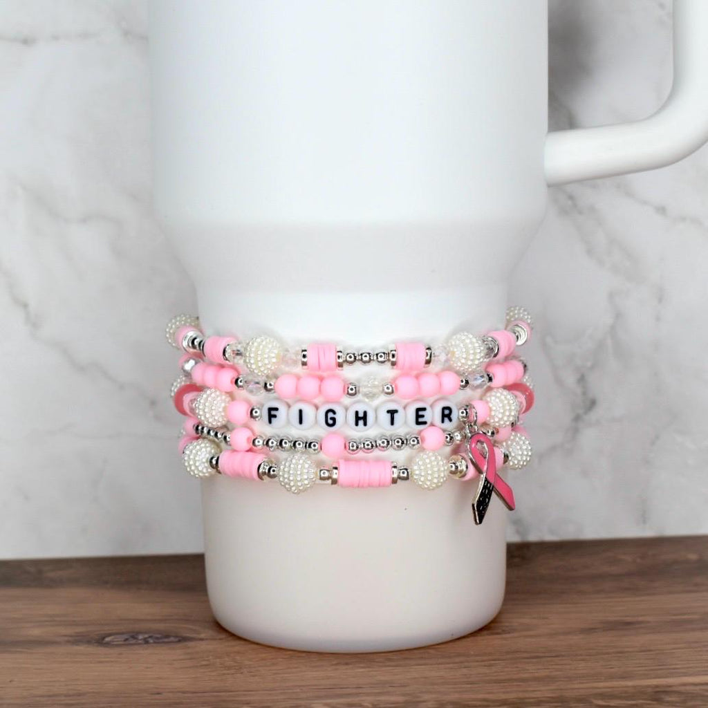 Stack of pink and white beaded bracelets with the word "FIGHTER" spelled out in letter beads. A pink ribbon charm hangs from one bracelet. The bracelets are displayed on a white tumbler.