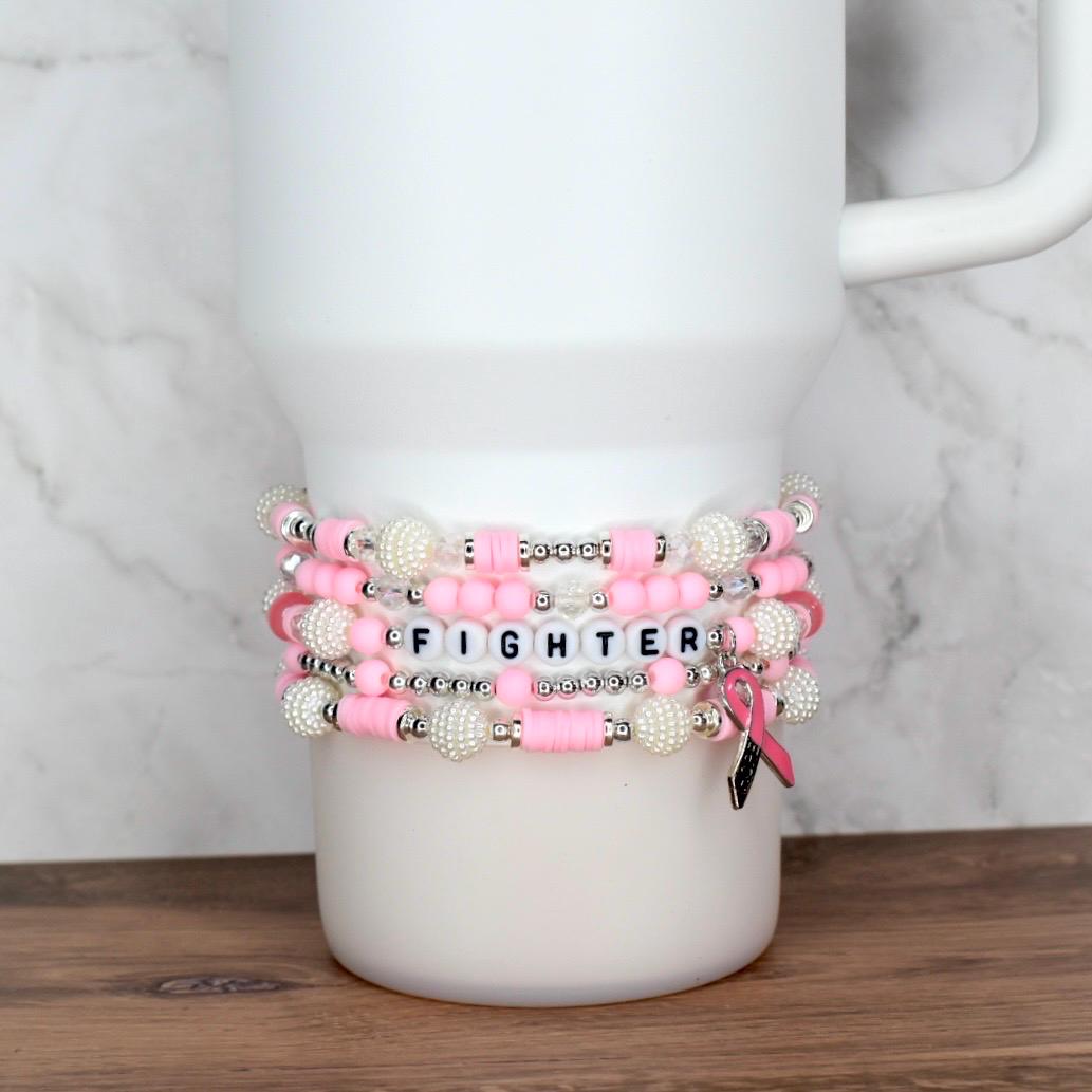 Stack of pink and white beaded bracelets with the word "FIGHTER" spelled out in letter beads. A pink ribbon charm hangs from one bracelet. The bracelets are displayed on a white tumbler.