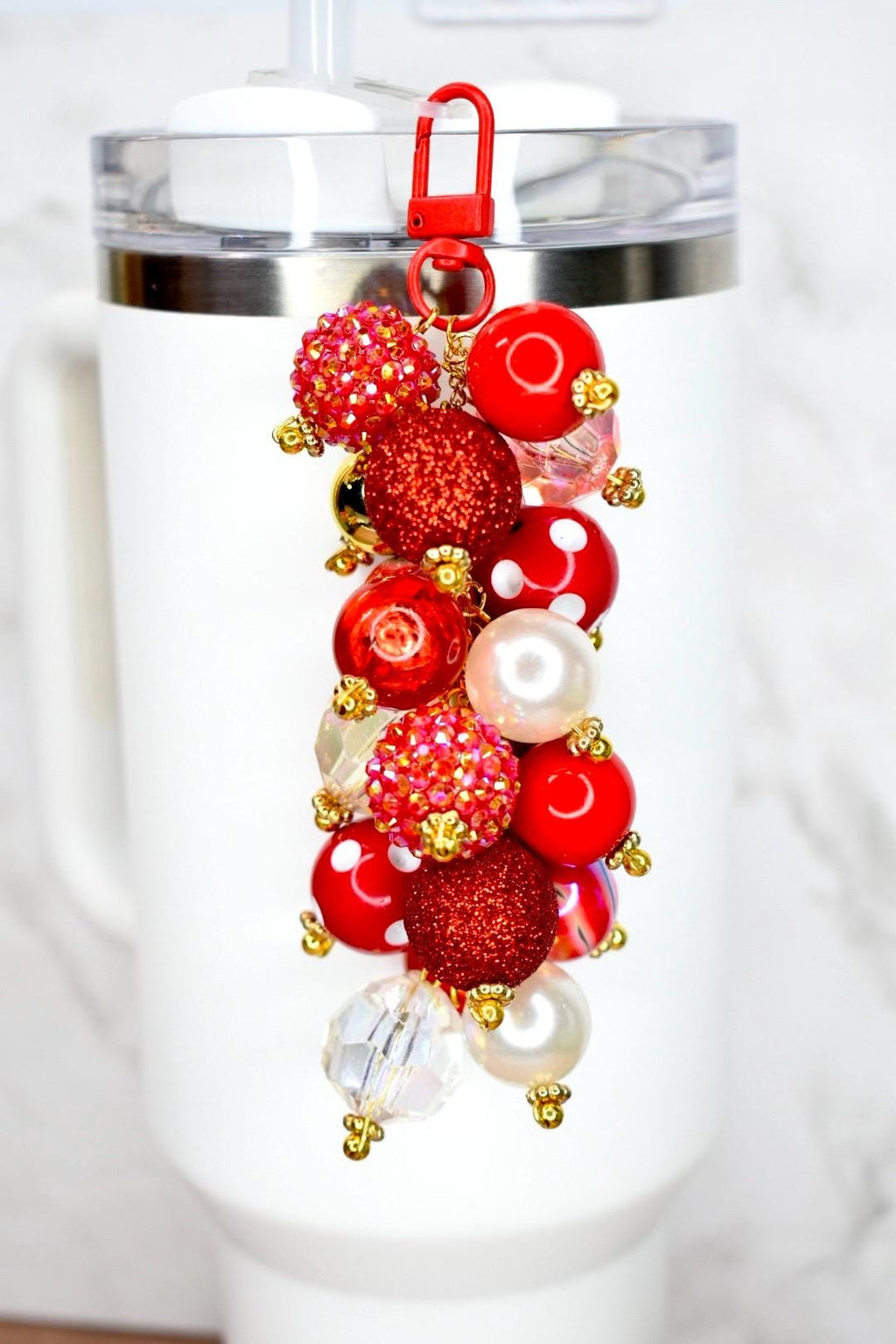 A tumbler bauble charm featuring a cluster of colorful bubblegum beads. The charm has a red lobster clasp and a gold chain. The beads are various sizes and colors, including red, gold, and clear. Some beads are glittery or have white polka dots. The charm is attached to a white tumbler with a straw.