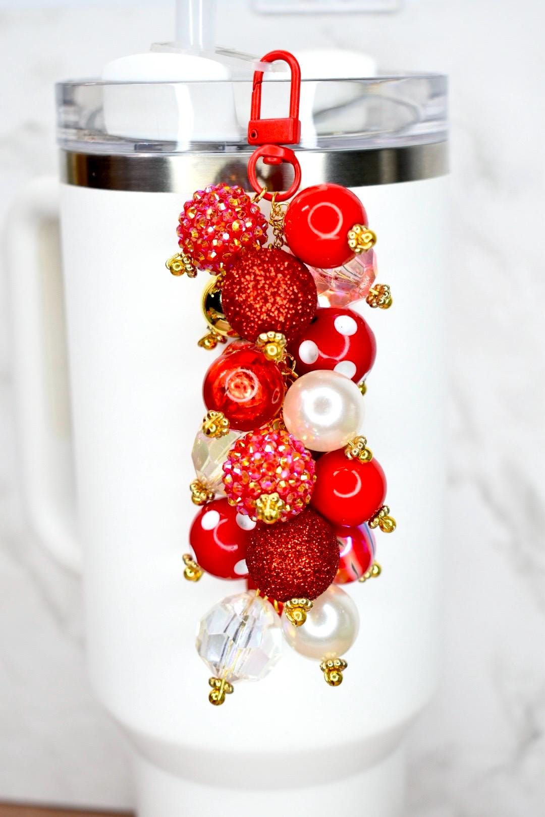 A tumbler bauble charm featuring a cluster of colorful bubblegum beads. The charm has a red lobster clasp and a gold chain. The beads are various sizes and colors, including red, gold, and clear. Some beads are glittery or have white polka dots. The charm is attached to a white tumbler with a straw.