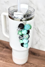 A white tumbler with a clear lid and straw, adorned with a beaded charm. The charm features a cluster of colorful beads in various patterns and textures, including turquoise, black, and green, attached with a silver clasp.