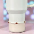 A white Stanley with a small bracelet wrapped around it. The bracelet features small white pearls, gold beads, and heart-shaped red gemstones. The mug has a handle and a smooth, matte finish.