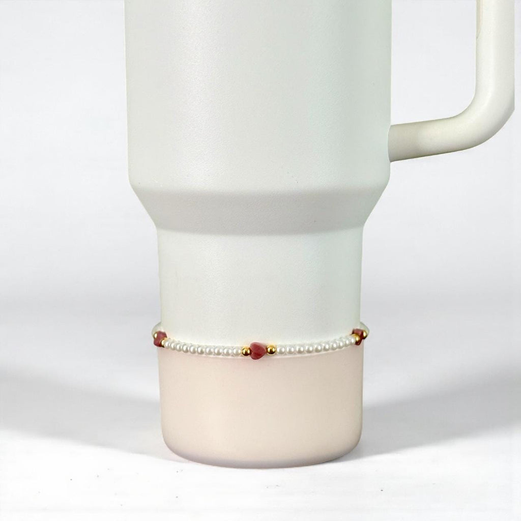 A white Stanley with a handle, featuring a delicate bracelet. The bracelet is composed of small white pearls, interspersed with red heart-shaped beads and gold accents. The bracelet is wrapped around the base of the tumbler.