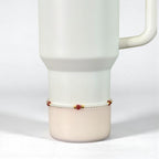 A white Stanley with a handle, featuring a delicate bracelet. The bracelet is composed of small white pearls, interspersed with red heart-shaped beads and gold accents. The bracelet is wrapped around the base of the tumbler.