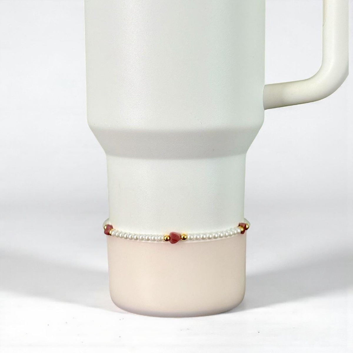 A white Stanley with a handle, featuring a delicate bracelet. The bracelet is composed of small white pearls, interspersed with red heart-shaped beads and gold accents. The bracelet is wrapped around the base of the tumbler.