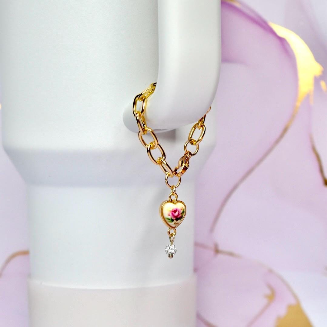 A gold-colored chain with a heart-shaped charm. The charm features a pink rose design and a small, clear crystal. The chain is attached to a white object.