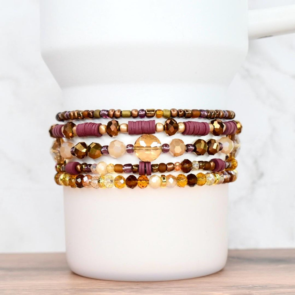 A collection of beaded bracelets in shades of gold, brown, and burgundy. The bracelets feature various bead sizes and shapes, including round, disc, and faceted beads. The bracelets are stacked on a white cylindrical object, showcasing the different designs.