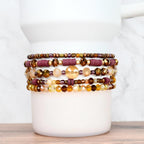 A collection of beaded bracelets in shades of gold, brown, and burgundy. The bracelets feature various bead sizes and shapes, including round, disc, and faceted beads. The bracelets are stacked on a white cylindrical object, showcasing the different designs.