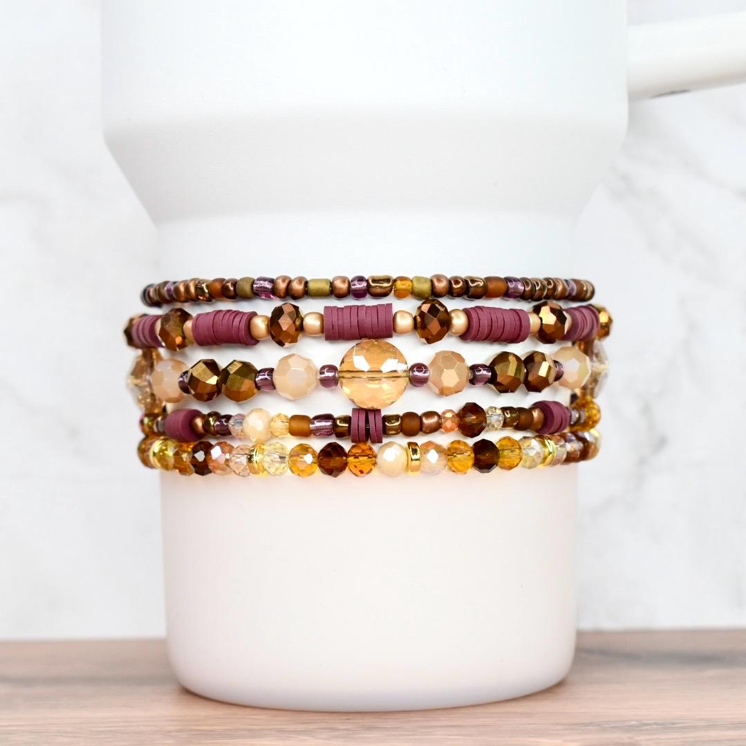 A collection of beaded bracelets in shades of gold, brown, and burgundy. The bracelets feature various bead sizes and shapes, including round, disc, and faceted beads. The bracelets are stacked on a white cylindrical object, showcasing the different designs.