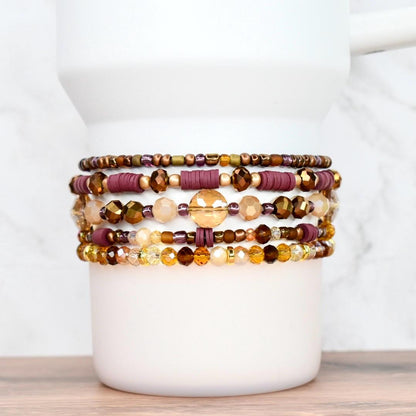 A collection of beaded bracelets in shades of gold, brown, and burgundy. The bracelets feature various bead sizes and shapes, including round, disc, and faceted beads. The bracelets are stacked on a white cylindrical object, showcasing the different designs.