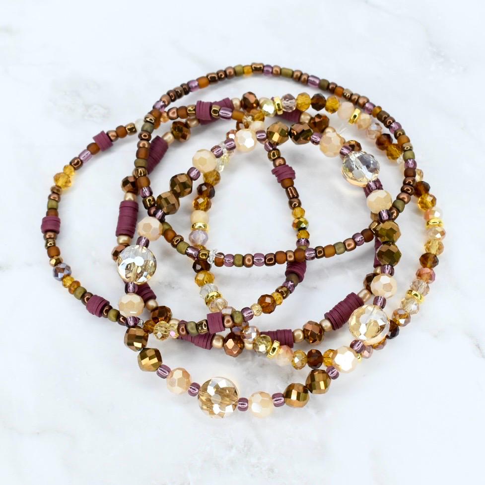 A collection of beaded bracelets in shades of brown, gold, and purple. The bracelets feature a variety of beads, including small seed beads, larger faceted beads, and cylindrical spacers. The bracelets are arranged in a circular pattern.