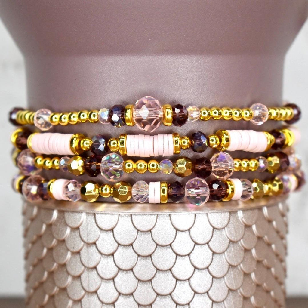Four stacked bracelets with a variety of beads. The bracelets feature gold-colored beads, pink disc beads, and faceted purple and clear beads. The bracelets are displayed on a textured, silver-colored surface.