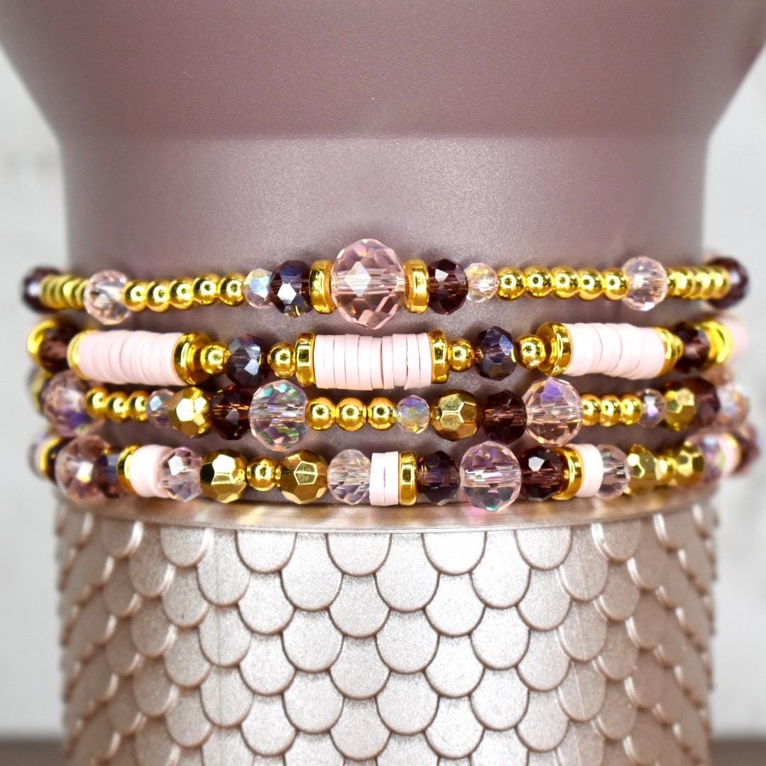 Four stacked bracelets with a variety of beads. The bracelets feature gold-colored beads, pink disc beads, and faceted purple and clear beads. The bracelets are displayed on a textured, silver-colored surface.