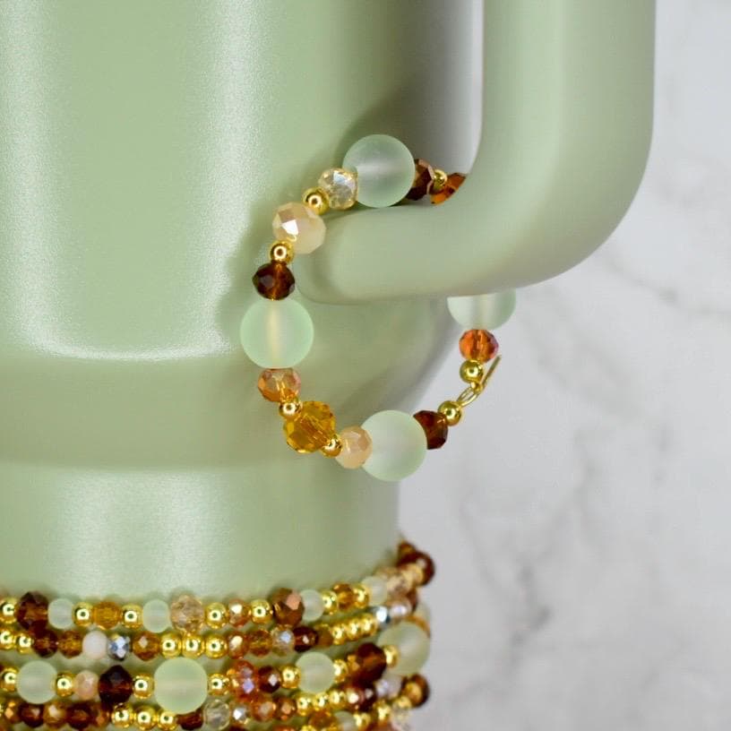 A beaded bracelet with a mix of colors, including light green, brown, gold, and peach. The bracelet is made with various sizes and shapes of beads, strung on a gold-colored wire. The bracelet is displayed on a light green ceramic object.
