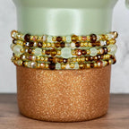 A stack of beaded bracelets in shades of gold, brown, and pale green. The bracelets are wrapped around a cylindrical, copper-colored container with a glittery finish. The background is a light green vase.