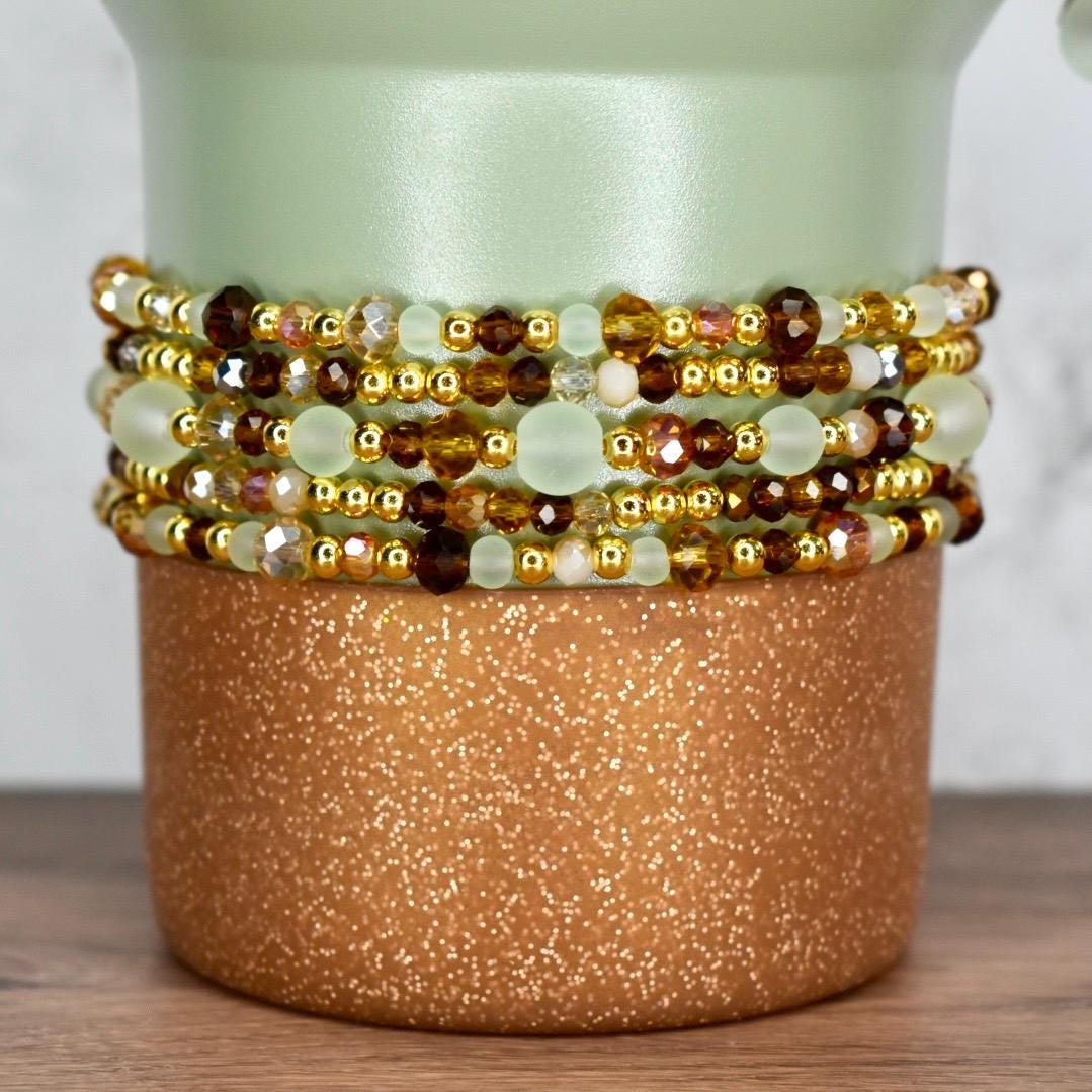 A stack of beaded bracelets in shades of gold, brown, and pale green. The bracelets are wrapped around a cylindrical, copper-colored container with a glittery finish. The background is a light green vase.