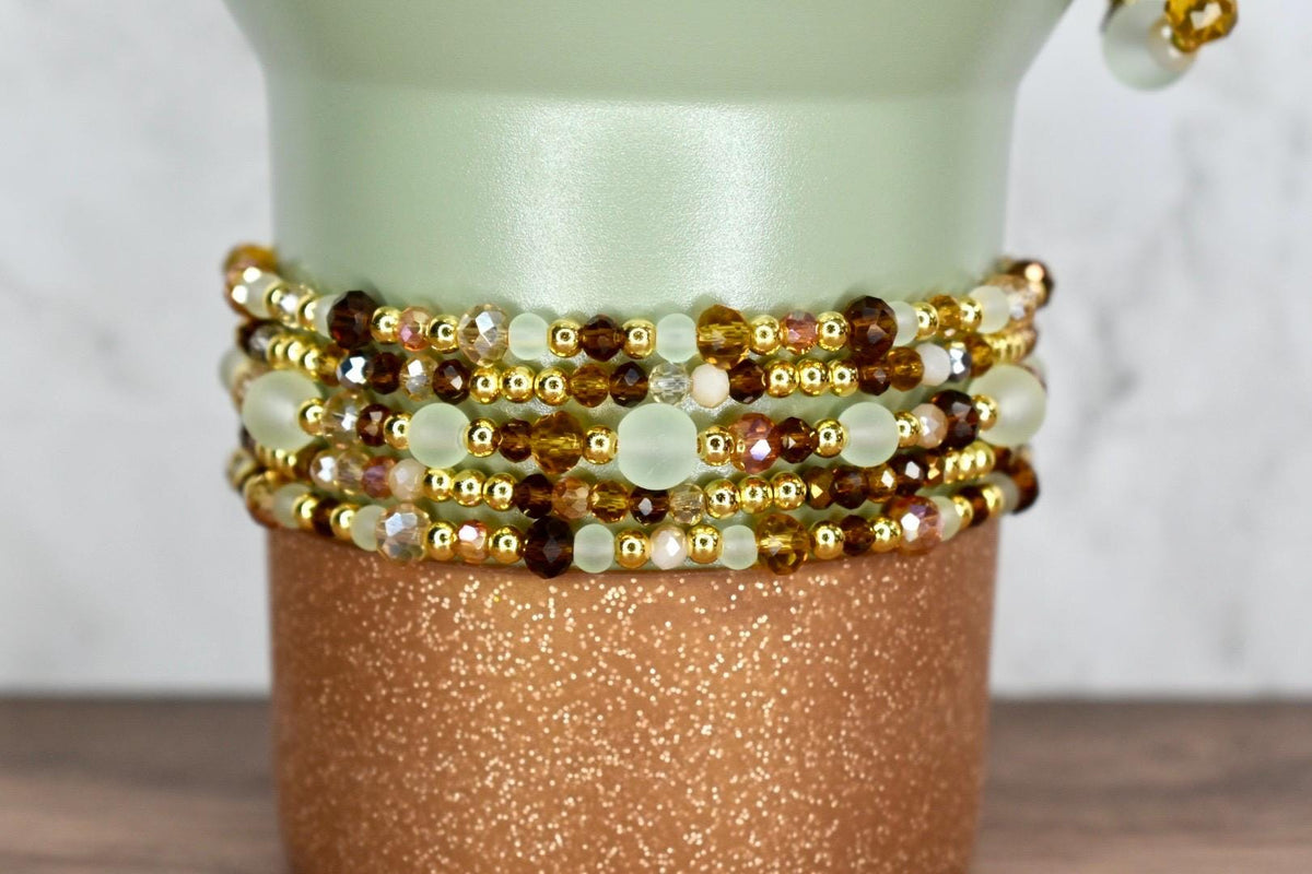 Close-up of a decorative vase with multiple beaded bracelets. The bracelets feature gold, brown, and clear beads. The vase is light green with a glittery copper-colored base.