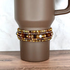 A brown travel mug with a beaded wrap. The wrap features multiple strands of amber, gold, and brown beads, with a few white beads interspersed. The mug has a handle and sits on a wooden surface.