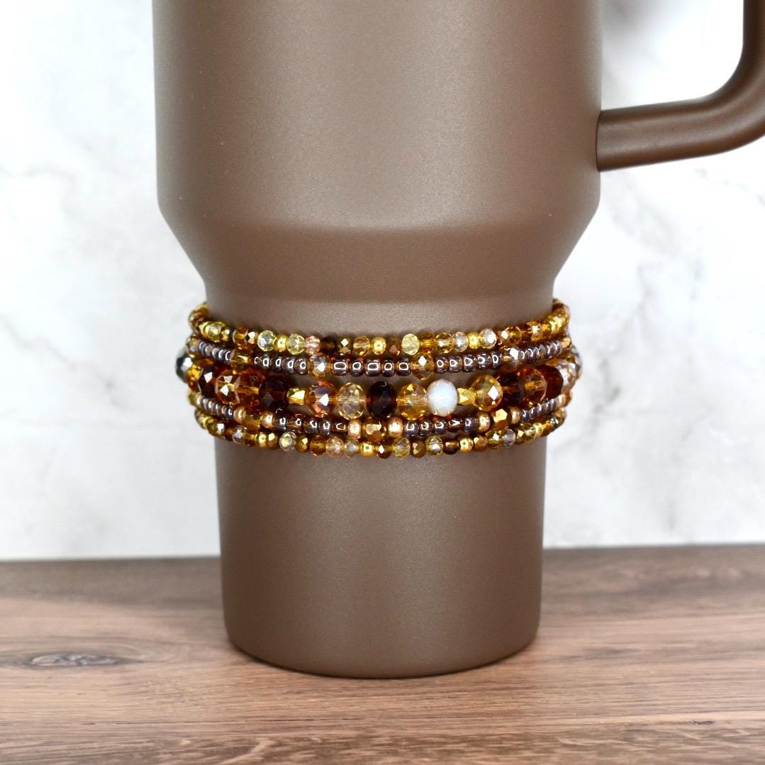 A brown travel mug with a beaded wrap. The wrap features multiple strands of amber, gold, and brown beads, with a few white beads interspersed. The mug has a handle and sits on a wooden surface.