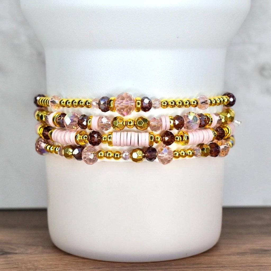 Stack of five beaded bracelets. The bracelets feature a mix of gold, pink, and purple beads. Some bracelets have gold-colored spacer beads, while others have pink disc beads. The bracelets are stacked on a white cylindrical object.
