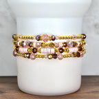 Stack of five beaded bracelets. The bracelets feature a mix of gold, pink, and purple beads. Some bracelets have gold-colored spacer beads, while others have pink disc beads. The bracelets are stacked on a white cylindrical object.