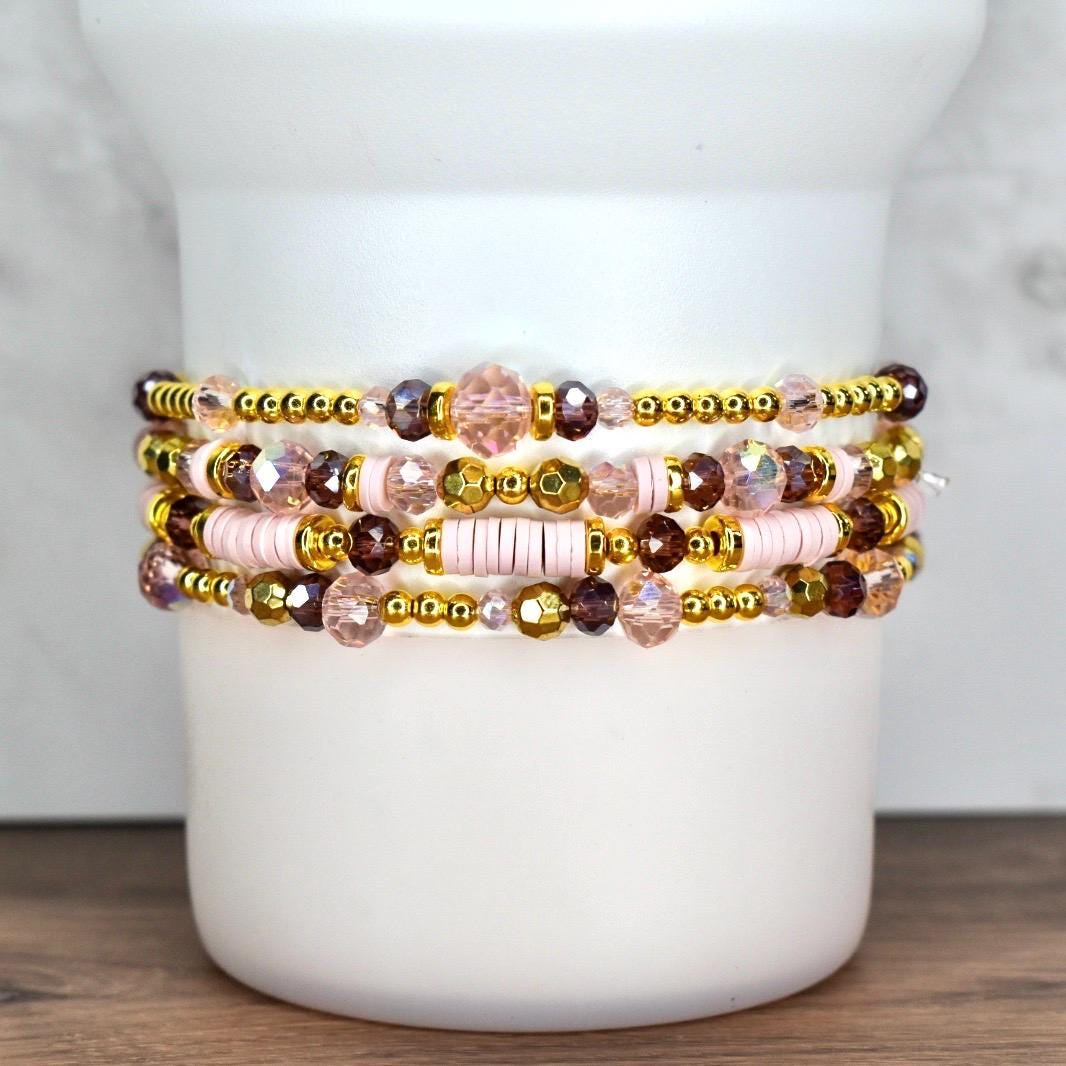 Stack of five beaded bracelets. The bracelets feature a mix of gold, pink, and purple beads. Some bracelets have gold-colored spacer beads, while others have pink disc beads. The bracelets are stacked on a white cylindrical object.