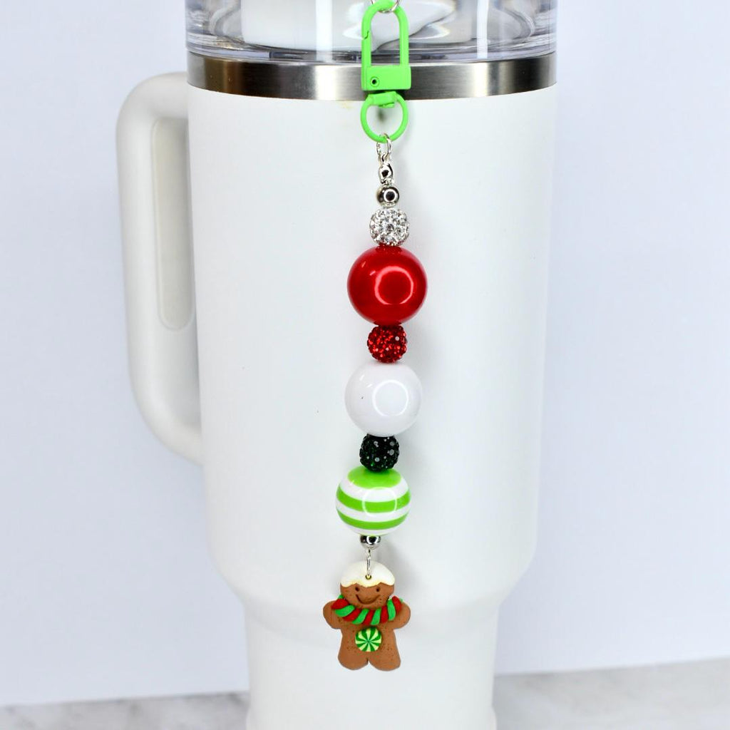 A Christmas-themed beaded straw charm with a green clip, attached to a white tumbler. The charm features red, white, and green beads, and a gingerbread man charm with a red and green scarf. The charm is designed to be attached to a tumbler.