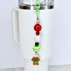 A Christmas-themed beaded straw charm with a green clip, attached to a white tumbler. The charm features red, white, and green beads, and a gingerbread man charm with a red and green scarf. The charm is designed to be attached to a tumbler.