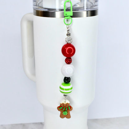 A Christmas-themed beaded straw charm with a green clip, attached to a white tumbler. The charm features red, white, and green beads, and a gingerbread man charm with a red and green scarf. The charm is designed to be attached to a tumbler.