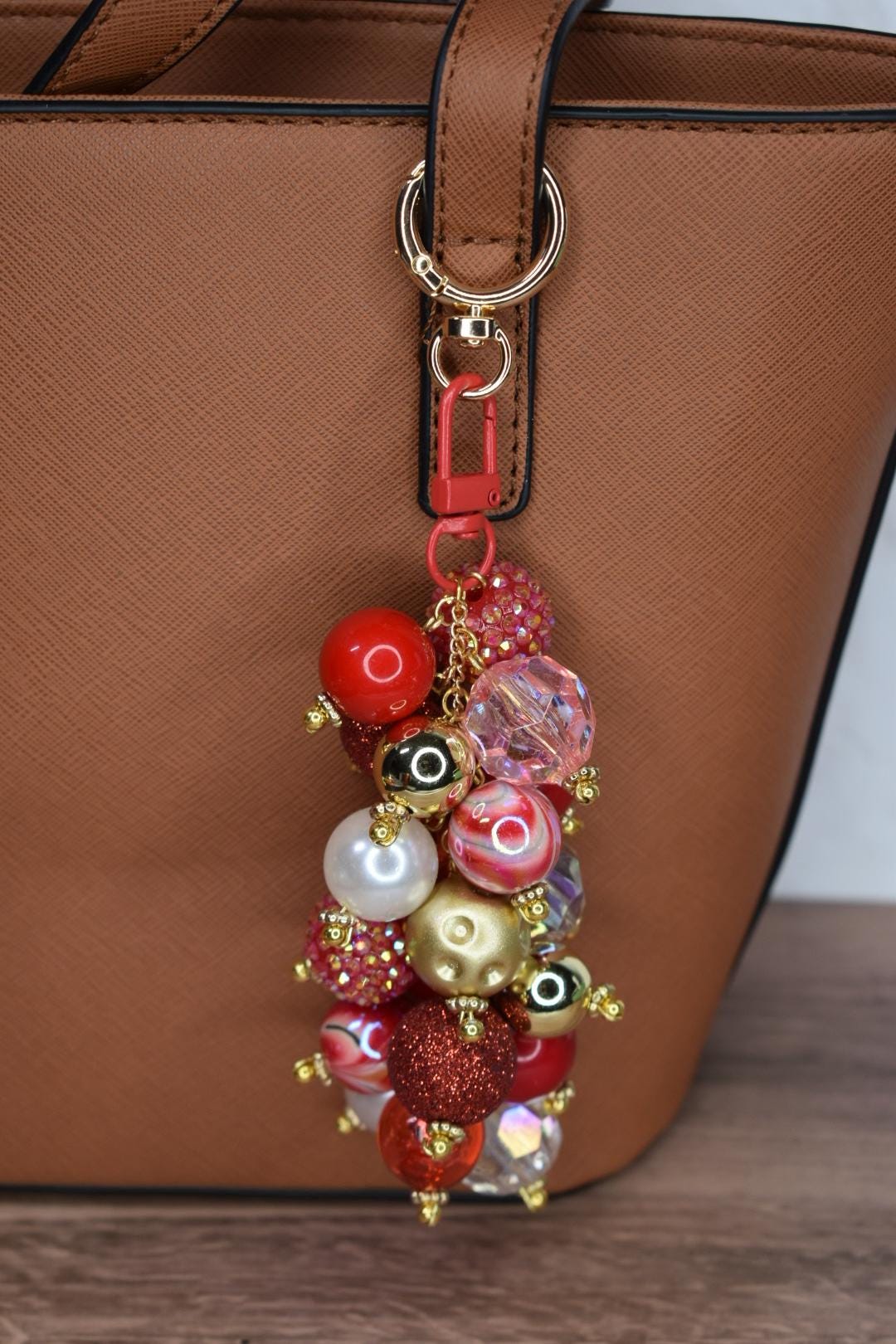 A beaded bag charm with a red lobster clasp and gold-tone hardware. The charm features a cluster of various sized beads in red, gold, pink, and clear colors. The charm is attached to a brown handbag.