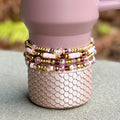 Stack of beaded bracelets in shades of pink, gold, and purple. The bracelets are made with various beads, including gold, iridescent, and round beads. The bracelets are stacked on a pink tumbler with a textured, scale-like pattern.