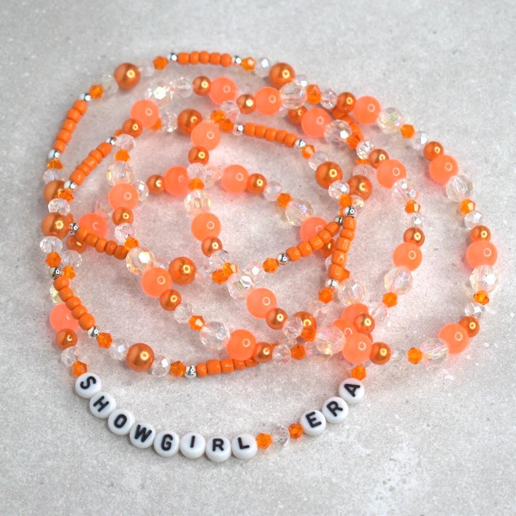 A bold bracelet stack in vibrant orange and crystal tones encircles a white tumbler, with “SHOWGIRL ERA” spelled out in chic white letter beads. The glowing orange background enhances the energetic, glamorous feel.