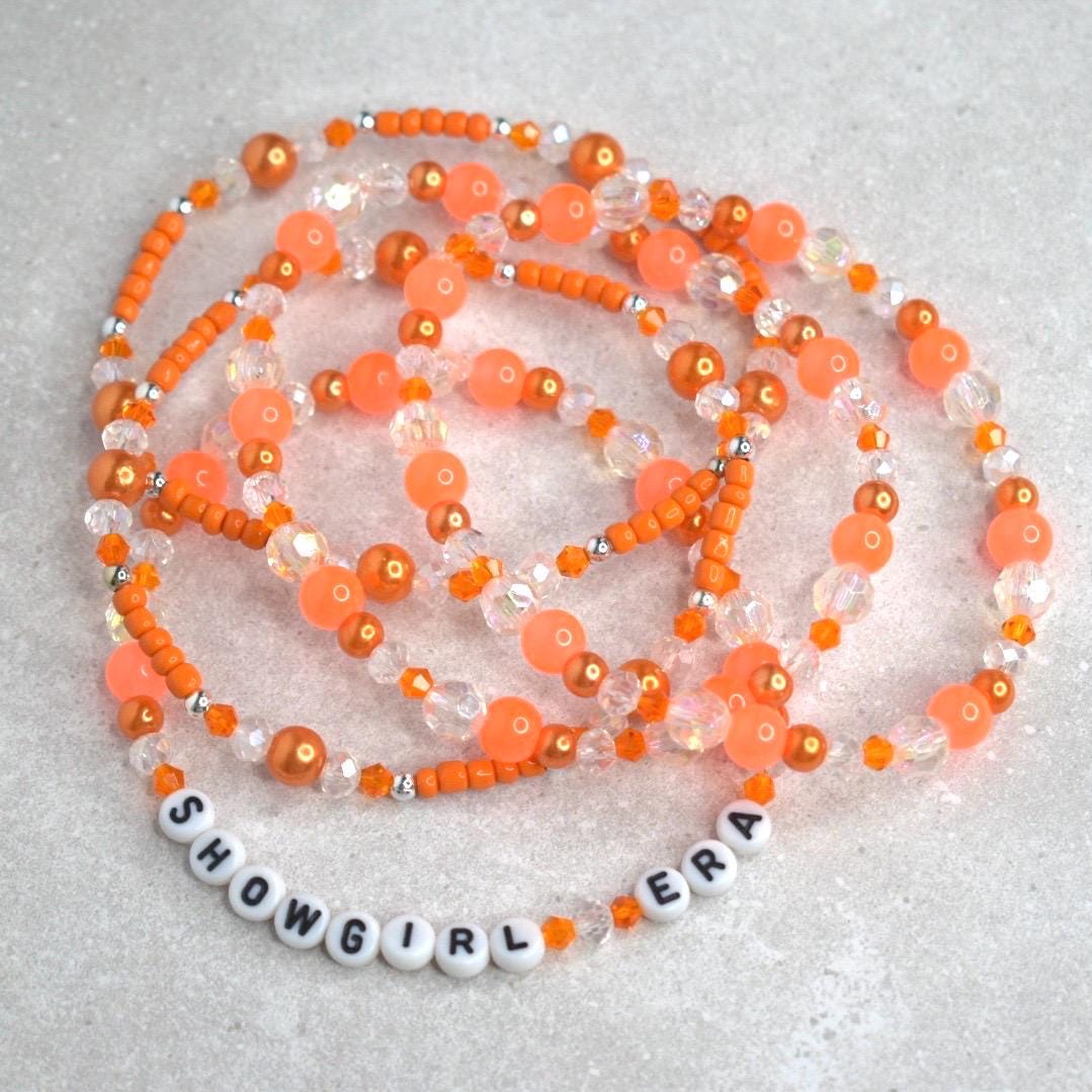 A bold bracelet stack in vibrant orange and crystal tones encircles a white tumbler, with “SHOWGIRL ERA” spelled out in chic white letter beads. The glowing orange background enhances the energetic, glamorous feel.