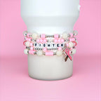 Stack of pink and white beaded bracelets with the word "FIGHTER" spelled out in letter beads. The bracelets have silver accents and a pink ribbon charm. The bracelets are displayed on a white cylindrical object against a pink background.