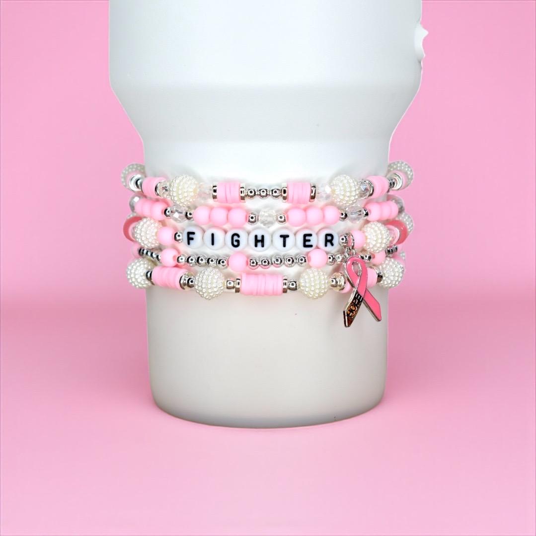 Stack of pink and white beaded bracelets with the word "FIGHTER" spelled out in letter beads. The bracelets have silver accents and a pink ribbon charm. The bracelets are displayed on a white cylindrical object against a pink background.