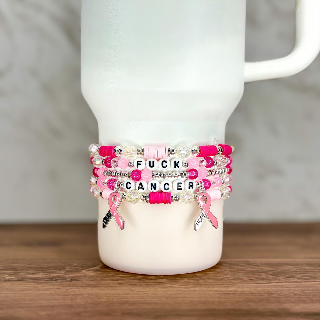 A stack of beaded bracelets in shades of pink and white, with the words "FUCK CANCER" spelled out in letter beads. Each bracelet has a pink ribbon charm. The bracelets are displayed on a white tumbler.