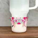 A stack of beaded bracelets in shades of pink and white, with the words "FUCK CANCER" spelled out in letter beads. Each bracelet has a pink ribbon charm. The bracelets are displayed on a white tumbler.