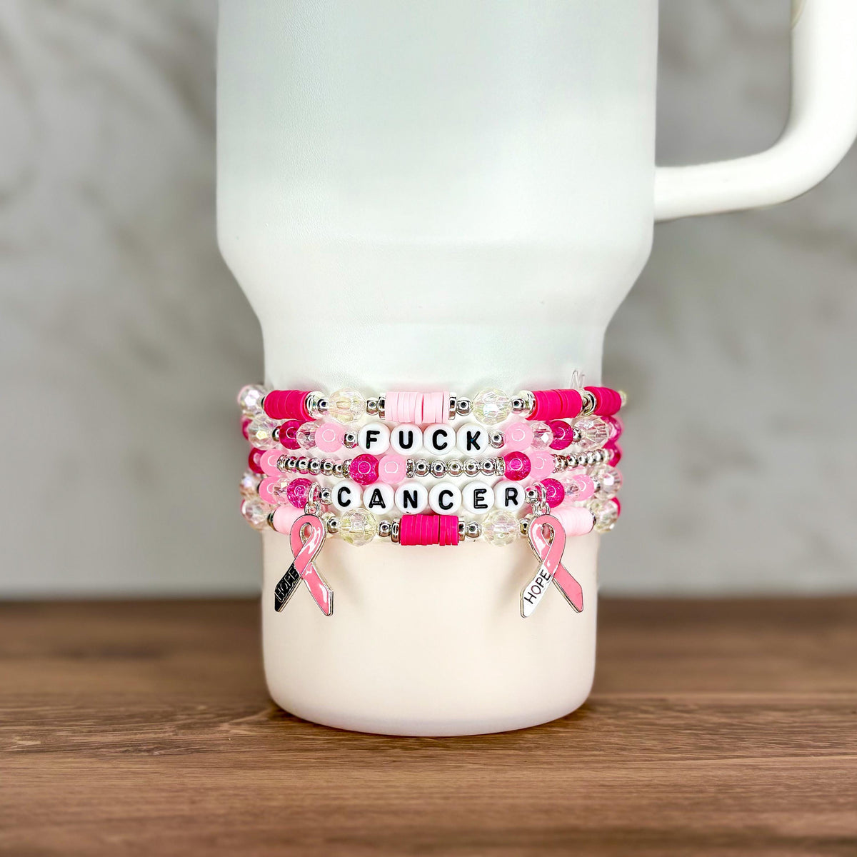 A stack of beaded bracelets in shades of pink and white, with the words "FUCK CANCER" spelled out in letter beads. Each bracelet has a pink ribbon charm. The bracelets are displayed on a white tumbler.