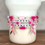 Stack of beaded bracelets in shades of pink, white, and clear. The bracelets have the words "FUCK CANCER" spelled out in white letter beads. Pink ribbon charms with the word "HOPE" hang from the bracelets.