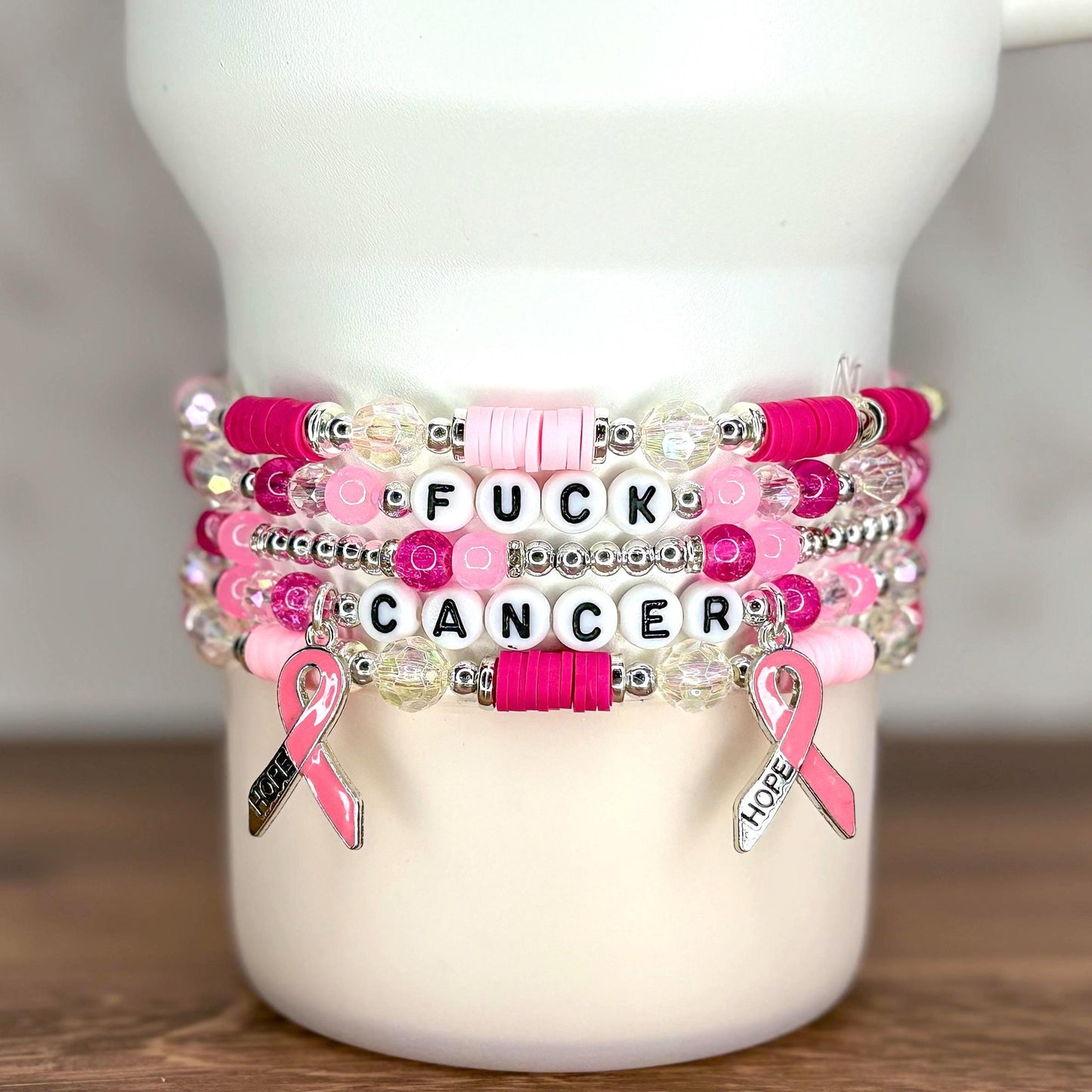 Stack of beaded bracelets in shades of pink, white, and clear. The bracelets have the words "FUCK CANCER" spelled out in white letter beads. Pink ribbon charms with the word "HOPE" hang from the bracelets.