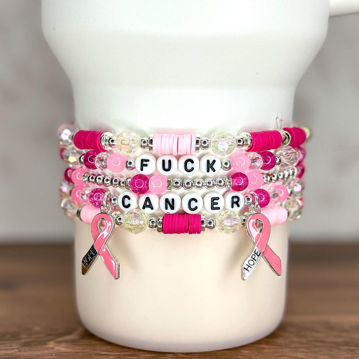 Stack of beaded bracelets in shades of pink, white, and clear. The bracelets have the words "FUCK CANCER" spelled out in white letter beads. Pink ribbon charms with the word "HOPE" hang from the bracelets.