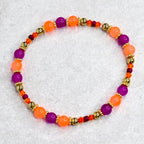 A beaded bracelet featuring alternating orange and purple beads, interspersed with gold-colored beads and smaller orange and red beads. The bracelet is strung on an elastic cord and is designed to be worn around the wrist.