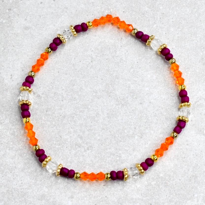 A beaded bracelet featuring a mix of orange, deep purple, and clear beads. Gold-colored accents separate the sections of beads. The bracelet is circular, and the beads are strung together to form a flexible piece of jewelry.