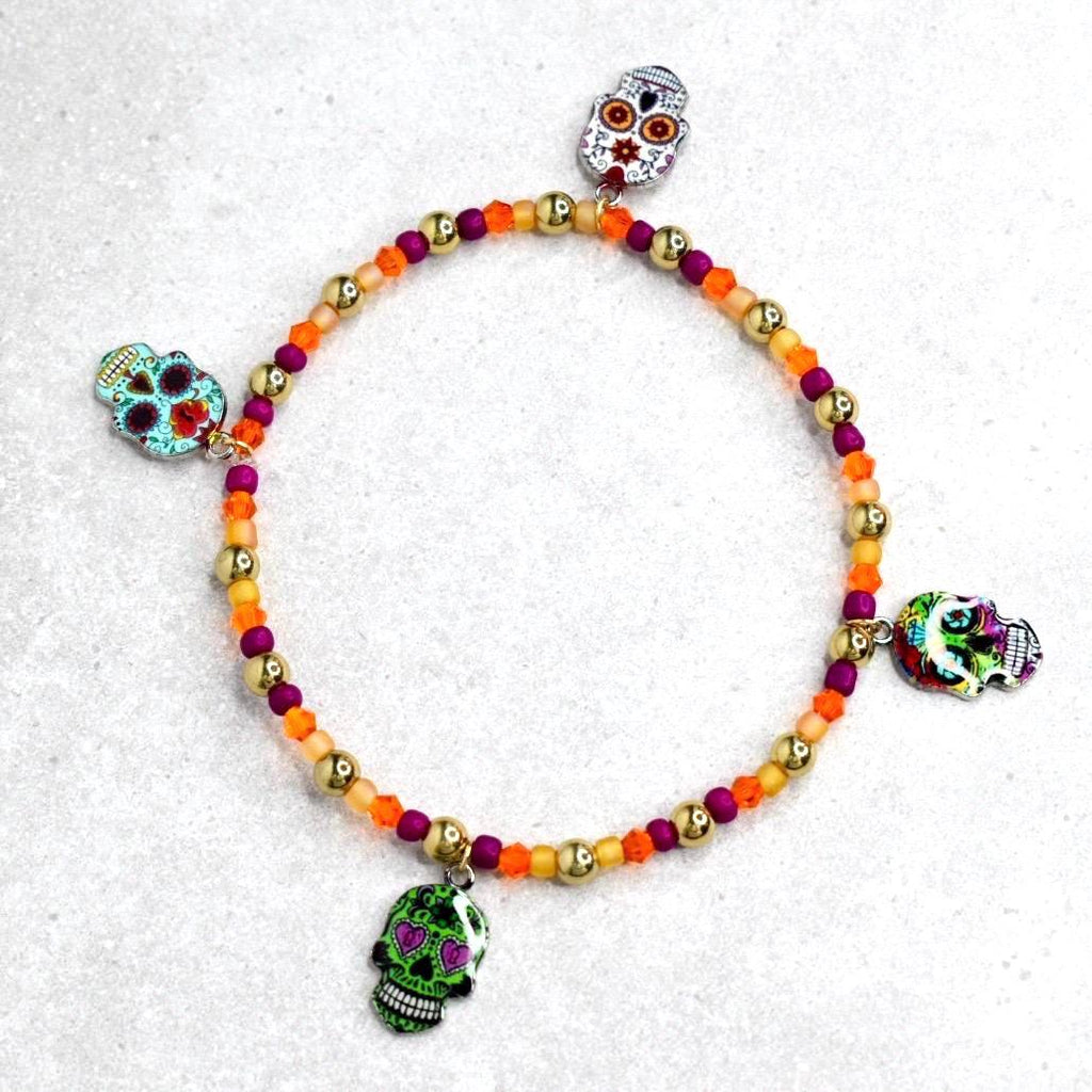 A beaded bracelet featuring a Day of the Dead theme. The bracelet has a mix of orange, gold, and purple beads. It is adorned with four skull charms in various colors and designs, adding a festive touch.