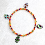 A beaded bracelet featuring a Day of the Dead theme. The bracelet has a mix of orange, gold, and purple beads. It is adorned with four skull charms in various colors and designs, adding a festive touch.