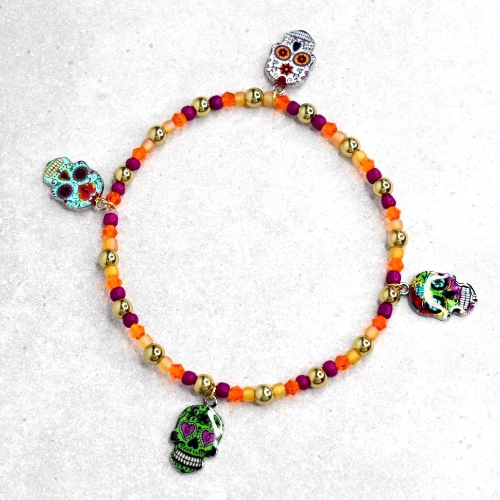 A beaded bracelet featuring a Day of the Dead theme. The bracelet has a mix of orange, gold, and purple beads. It is adorned with four skull charms in various colors and designs, adding a festive touch.