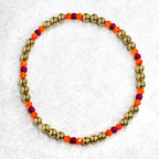 A gold beaded bracelet with alternating sections of gold beads, orange beads, and purple beads. The bracelet is circular and sits on a light gray surface. The beads are small and evenly spaced.