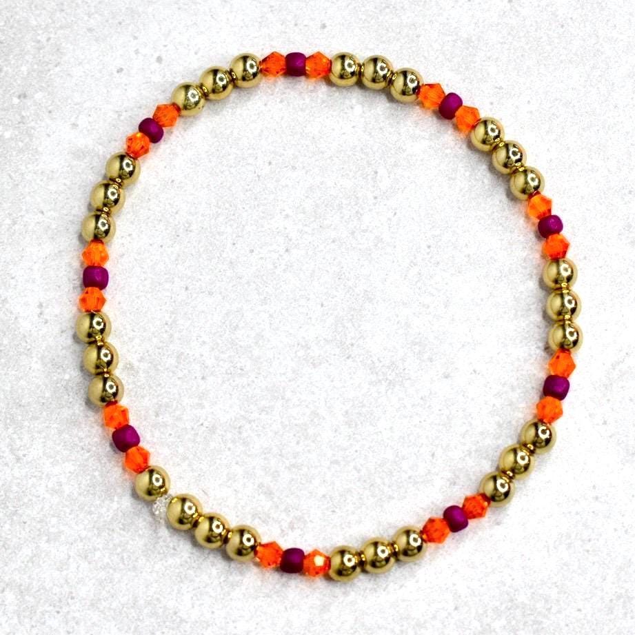 A gold beaded bracelet with alternating sections of gold beads, orange beads, and purple beads. The bracelet is circular and sits on a light gray surface. The beads are small and evenly spaced.