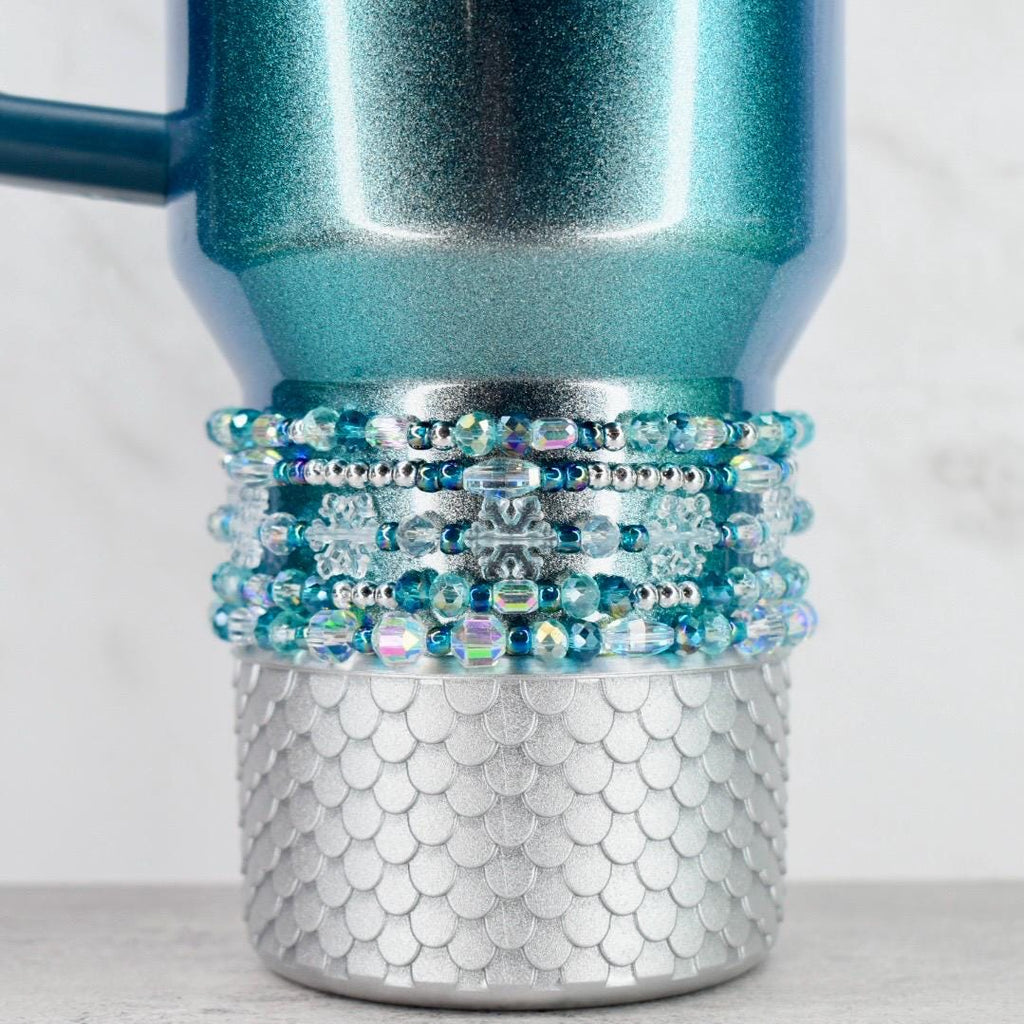 A teal and silver tumbler with a beaded accent. The upper portion of the tumbler is teal with a glittery finish. The middle section features multiple rows of blue and clear beads. The bottom has a silver, scale-like design.