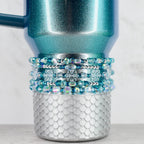 A teal and silver tumbler with a beaded accent. The upper portion of the tumbler is teal with a glittery finish. The middle section features multiple rows of blue and clear beads. The bottom has a silver, scale-like design.